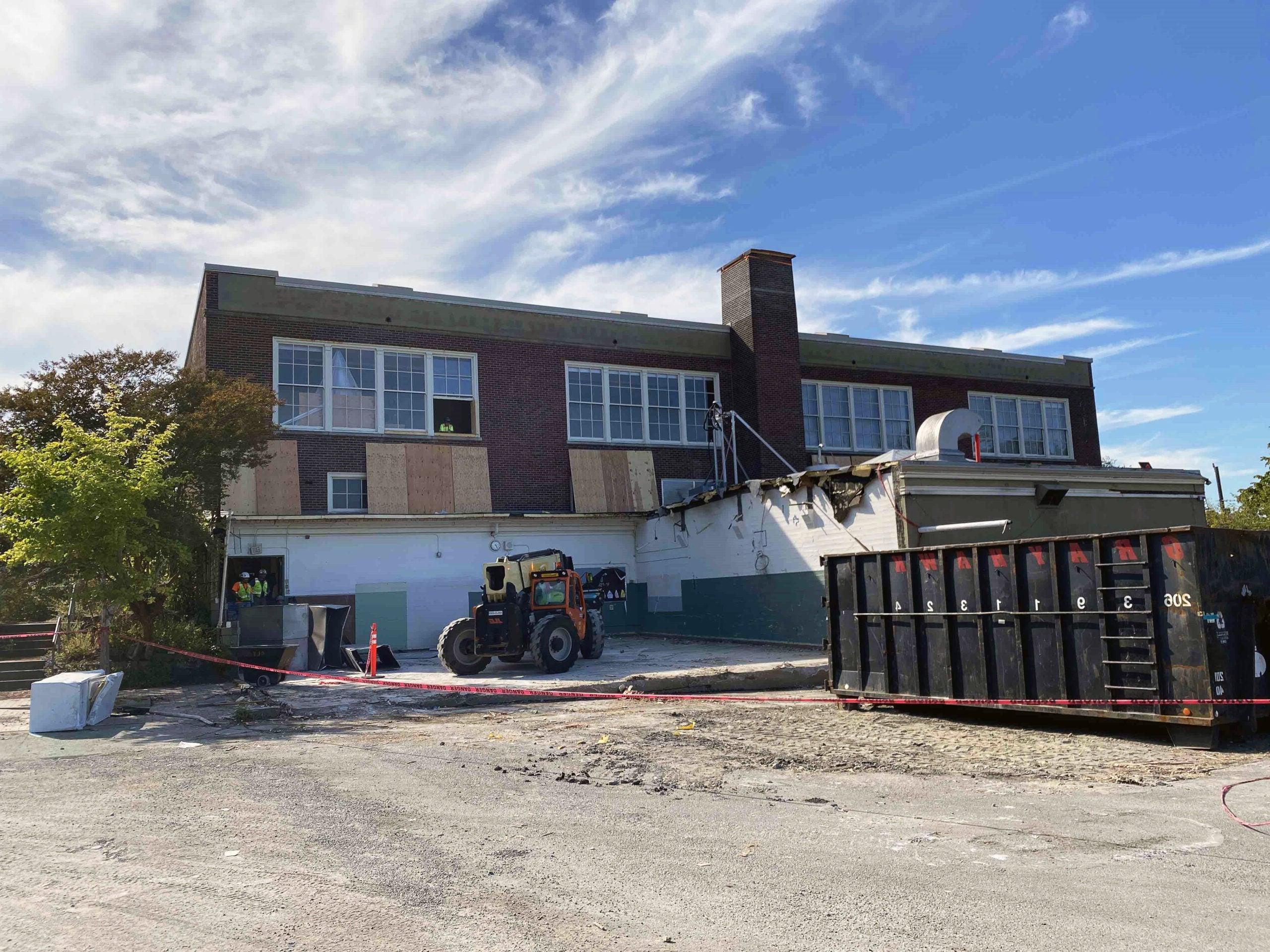 a two story brick building has windows on the top floor. an excavator is sitting below it on a concrete pad with only two walls remaining. a dumpster is next to the concrete